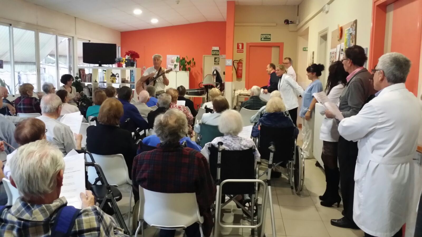 Vista general del salón: Joan Marc toca al fondo mientras un amplio grupo de personas mayores, algunas en silla de ruedas, escucha junto al personal del centro.