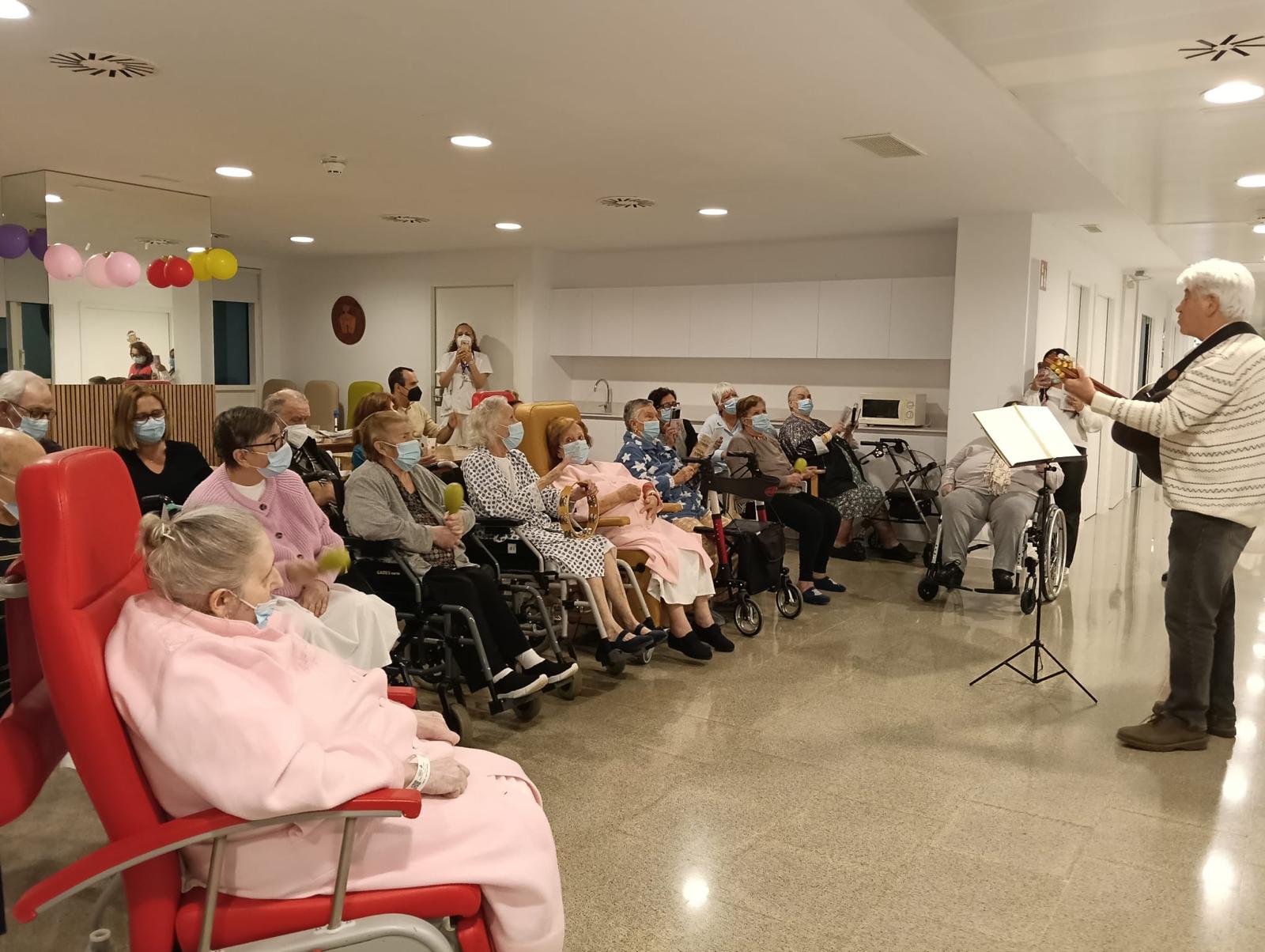 Joan Marc, de pie con jersey claro y guitarra, canta ante un grupo de personas sentadas en una sala del hospital, algunas acompañando con instrumentos de percusión.