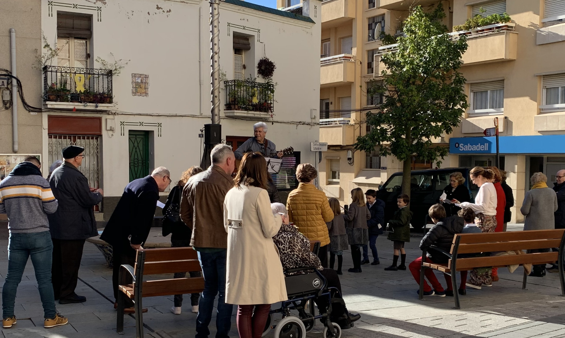 Joan Marc tocando la guitarra en una plaza soleada, rodeado de vecinos y niños que escuchan entre bancos y fachadas con balcones.