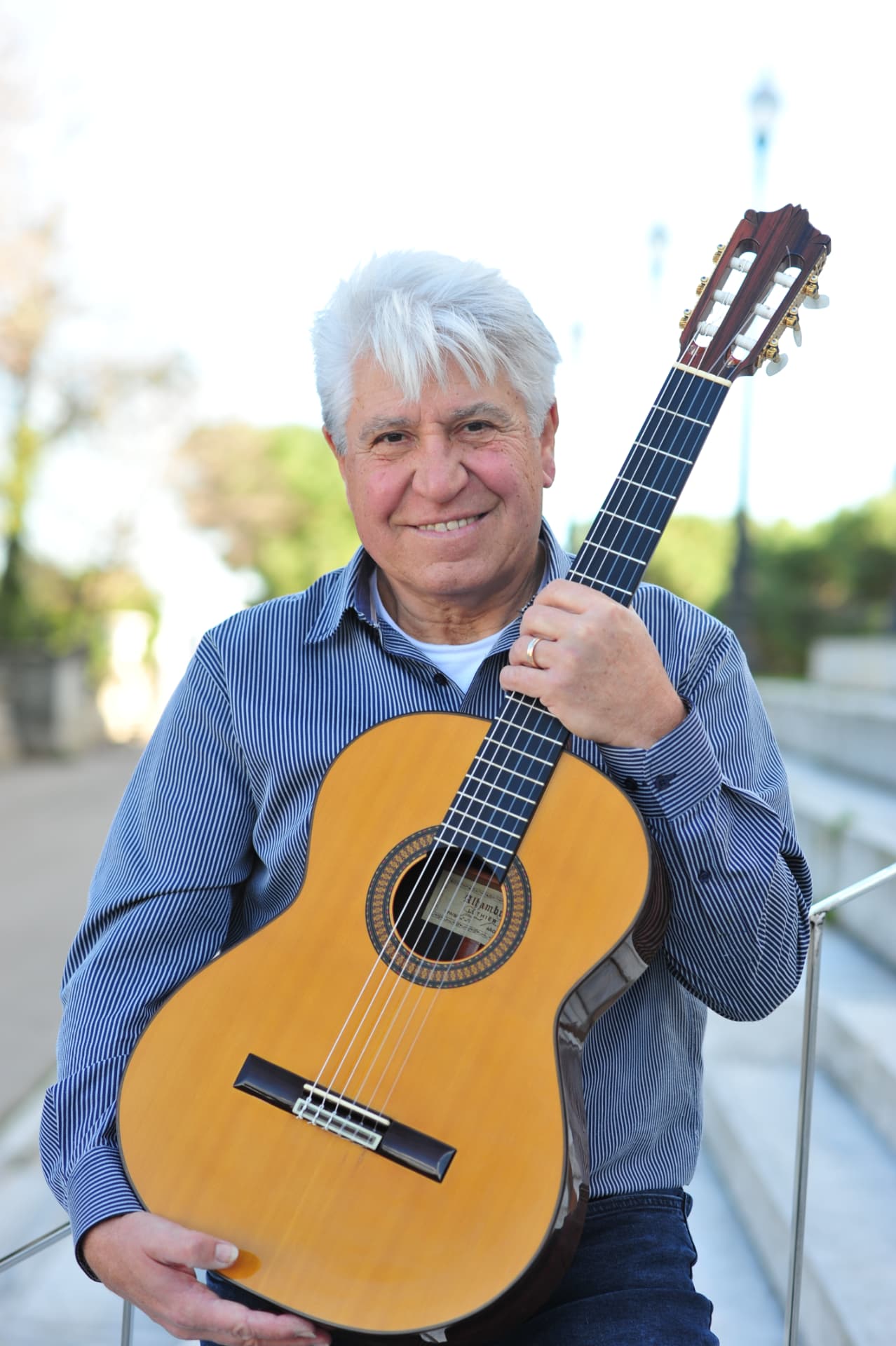 Joan Marc sonríe de frente, con camisa de rayas azules y la guitarra clásica abrazada al pecho, junto a una barandilla al aire libre.